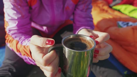 girl with polished red nails holding a cup of morning coffee in her hands while sitting on the sleeping bag