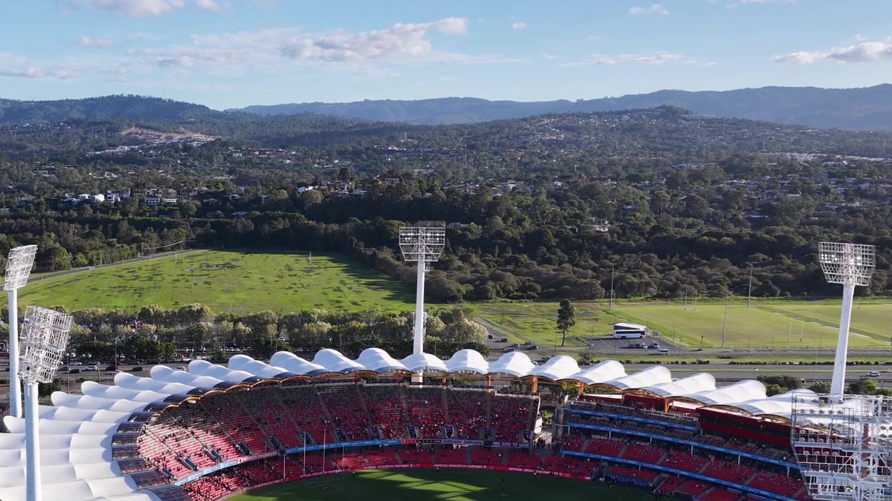 Drone pans across a large sports stadium with floodlights against a lush green mountain backdrop.