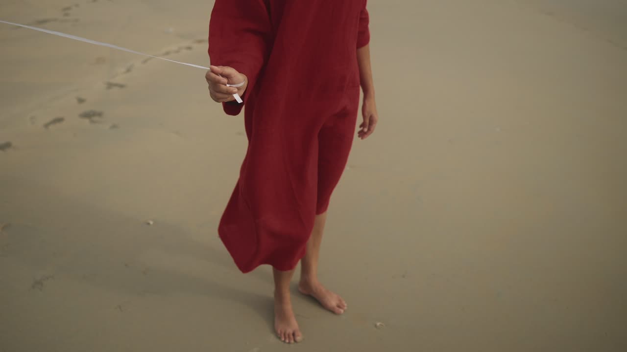 на пляже, одна и беззаботная, девушка босиком и в длинном красном топе держит веревку змея