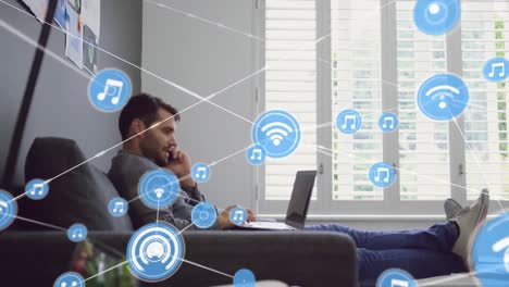 animación de iconos conectados, hombre caucásico sentado en el sofá y hablando por teléfono mientras usa una computadora portátil