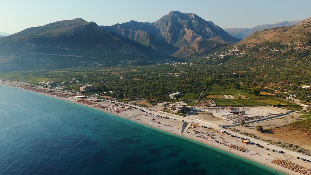 A serene beach with clear green water, scattered tourists, and mountains in the background, aerial view