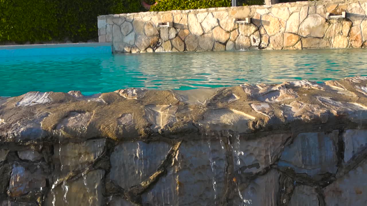 Luksushotel med swimmingpool i Portugal med turkisblåt vand, der flyder over stenkanten på et solrigt feriested, filmet i en langsom og glidende bevægelse