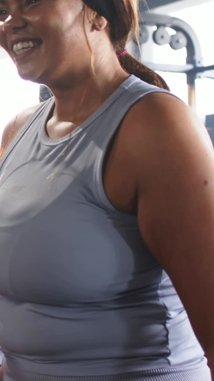 Video verticale: Zoom indietro che mostra una donna che risponde e si sistema in palestra, con rastrelliera per pesi, canotta e fascia per capelli.