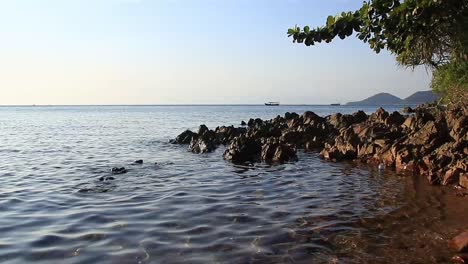 Hypnotisch-Plätschernde-Wellen-Am-Felsigen-Strand-Von-Koh-Tonsay-Oder-Rabbit-Island-In-Kambodscha-Mit-Einem-Vorbeifahrenden-Boot-Am-Horizont-Und-Einem-Ablenkenden-Plastikbecher,-Der-In-Der-Nähe-Schwimmt-Und-Das-Ausmaß-Der-Verschmutzung-Zeigt