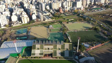 Vista-Aérea-Alejándose-De-Las-Canchas-De-Fútbol-Con-Sus-Jugadores-Y-Los-Edificios-De-La-Ciudad-Al-Fondo