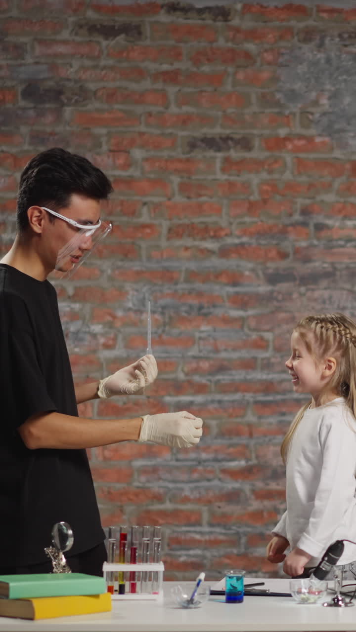 Little blonde girl student punches fist of positive Asian teacher holding material sample for experiment at fun science lesson in classroom