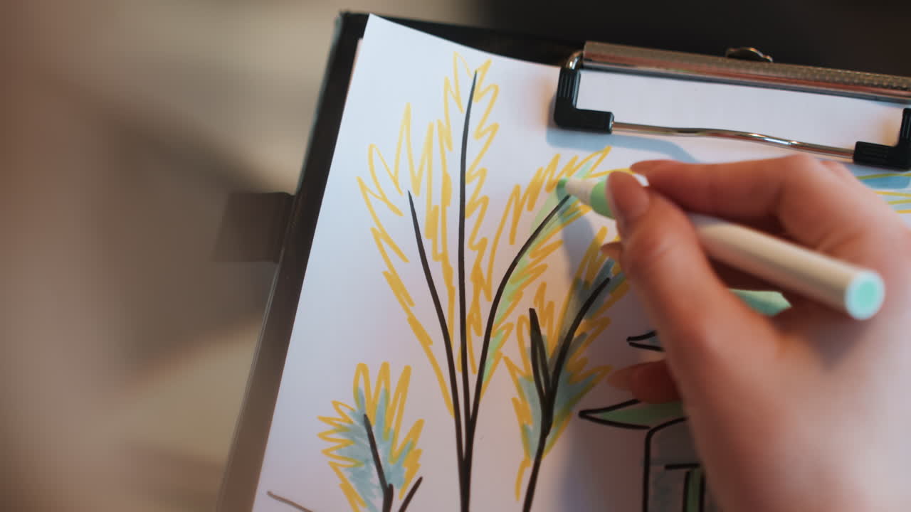 Primo piano di un artista bianco che disegna steli frondosi con pennarello verde e giallo su una tavoletta, sfondo sfocato, movimenti delicati delle mani, luce solare soffusa, toni naturali, atmosfera da sessione di studio, osservazionale