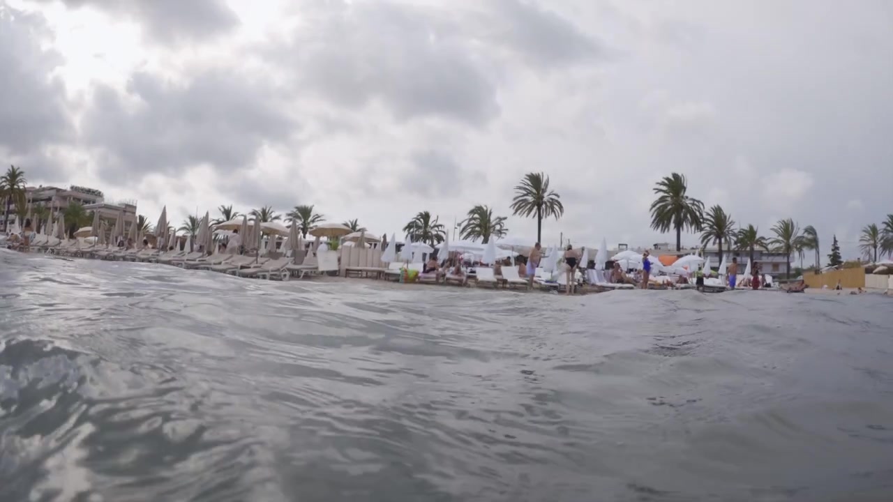Nyder en strandferie fra vandets perspektiv, der viser folk, der slapper af på liggestole og svømmer under en overskyet himmel.