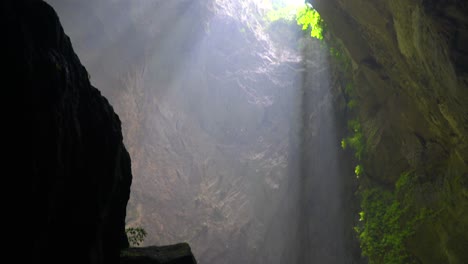 pan downward from the doline with sunbeam shining down to a explorer walking inside a dark cave