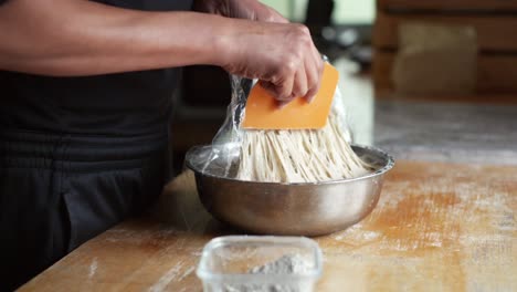 Puffy-and-sticky-dough-scraped-off-plastic-wrap-in-metal-bowl-using-orange-scraper,-filmed-as-close-up-slow-motion-shot