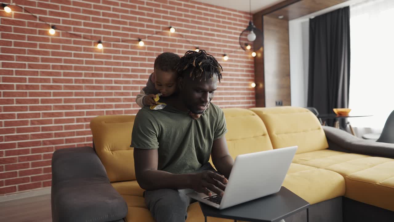 Smiling african father doing freelance work on laptop while his son disturbing him from the back