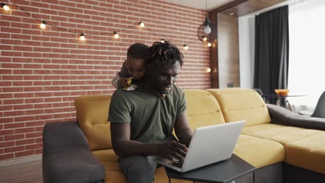 smiling african father doing freelance work on laptop while his son disturbing him from the back