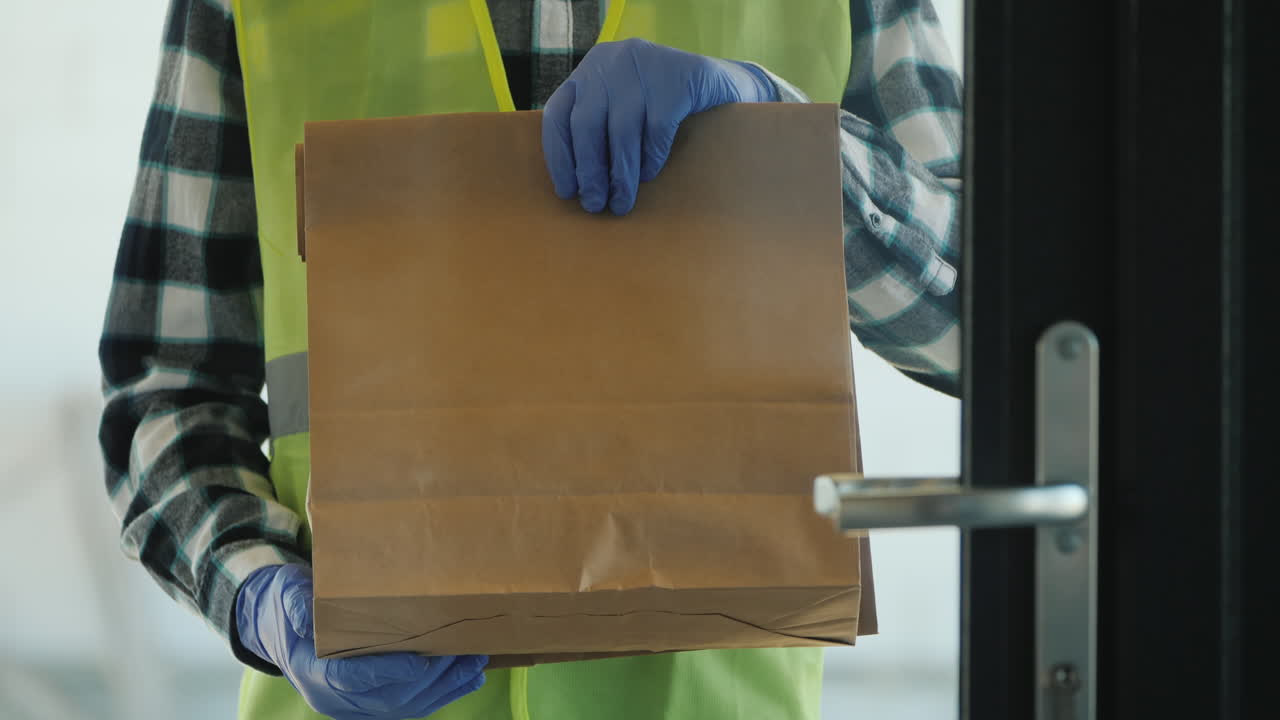 A Courier In Gloves With A Packet Of Groceries Stands Outside The Door
