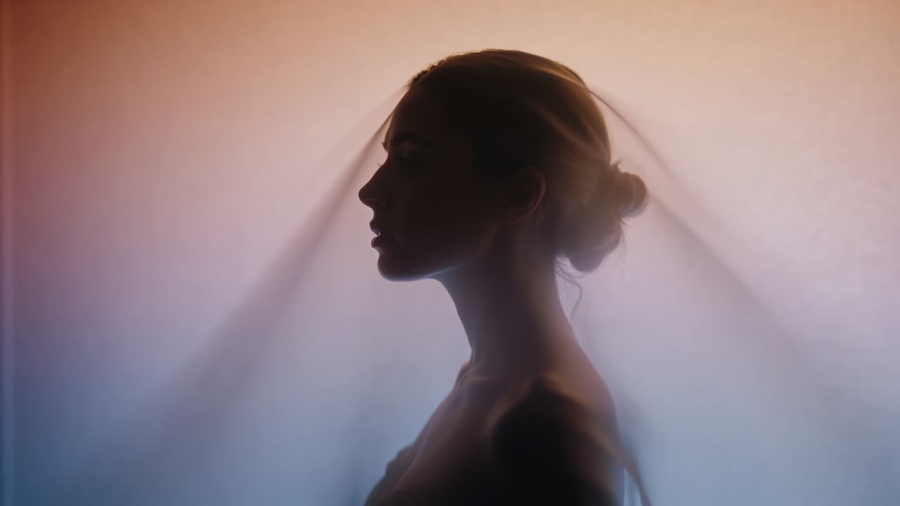 Inquadratura di una donna in profilo sinistro in un set retroilluminato, con il velo che si sposta all'indietro rivelando uno chignon basso e un abito.