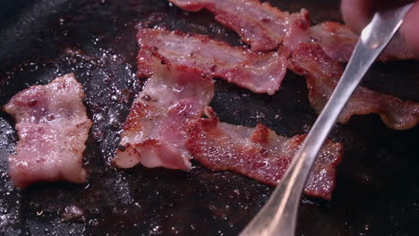 Tasty-bacon-sizzles-in-hot-frying-pan-as-fork-flips-strips,-closeup