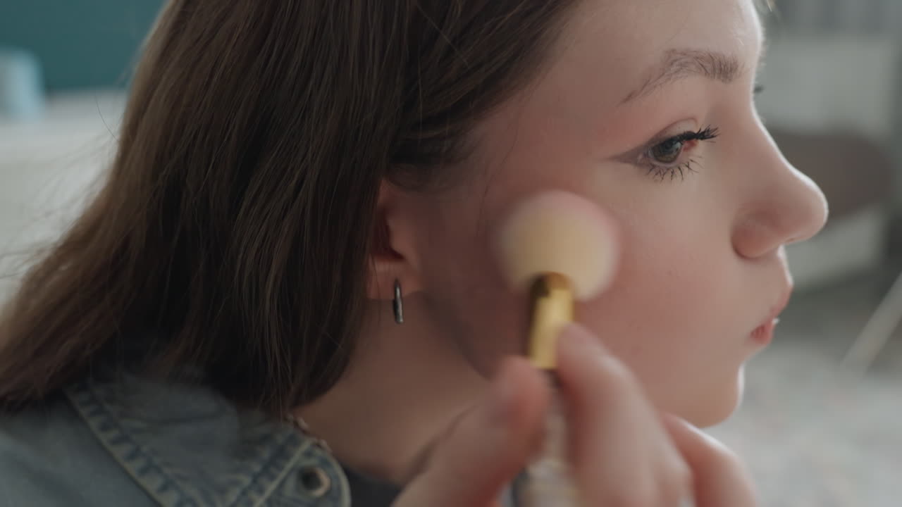 White teenage girl applying blush closeup, denim shirt, soft natural light, focused expression, precise brush strokes on cheek, subtle rosy glow, visible skin texture, intimate bedroom setting