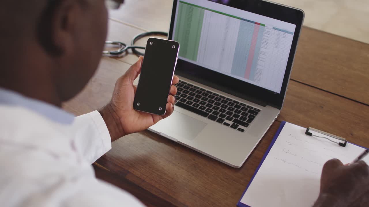 médico masculino afroamericano senior haciendo papeleo en una computadora portátil y usando un teléfono inteligente
