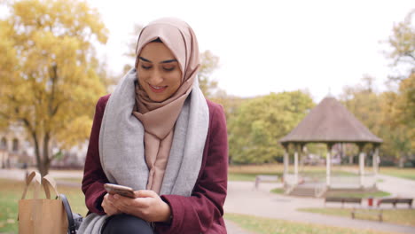 Mujer-Musulmana-Británica-Enviando-Mensajes-De-Texto-Por-Teléfono-Móvil-En-El-Parque