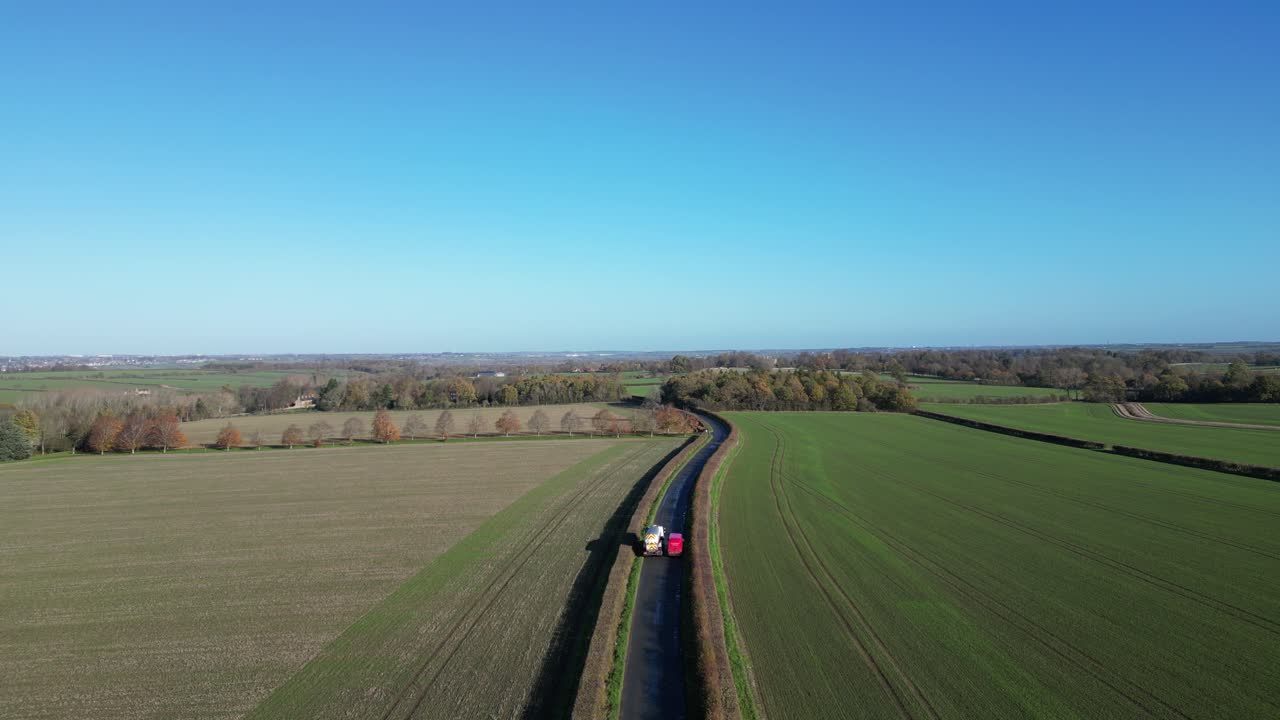 Drone shot of a winding rural road cutting through green farmland on a clear sunny day. Trees, fields, and a vibrant blue sky create a peaceful countryside scene.