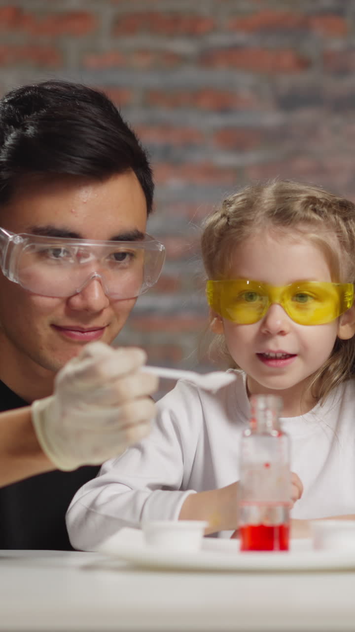 Happy little girl student nods looking at bottle of liquids while Asian lab assistant adds reagent with African-American colleague in studio