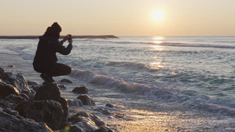 Der-Reisende-Nutzt-Sein-Telefon,-Um-Den-Morgendlichen-Blick-Auf-Das-Meer-Während-Des-Atemberaubenden-Sonnenaufgangs-Einzufangen,-Während-Sich-Die-Sonne-Auf-Der-Oberfläche-Spiegelt