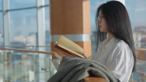 side view of focused student with long black hair reading book while leaning on wooden rail, coat draped beside her, sunlight streaming in through large windows with blurred cityscape in background