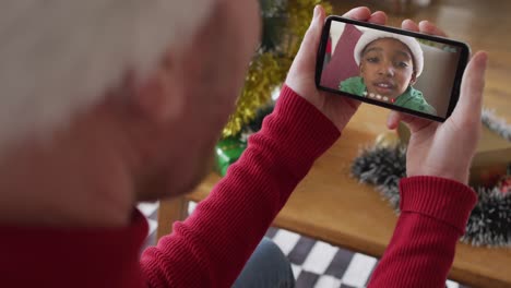 Hombre-Caucásico-Con-Sombrero-De-Santa-Usando-Un-Teléfono-Inteligente-Para-Una-Videollamada-Navideña-Con-Un-Niño-Sonriente-En-La-Pantalla