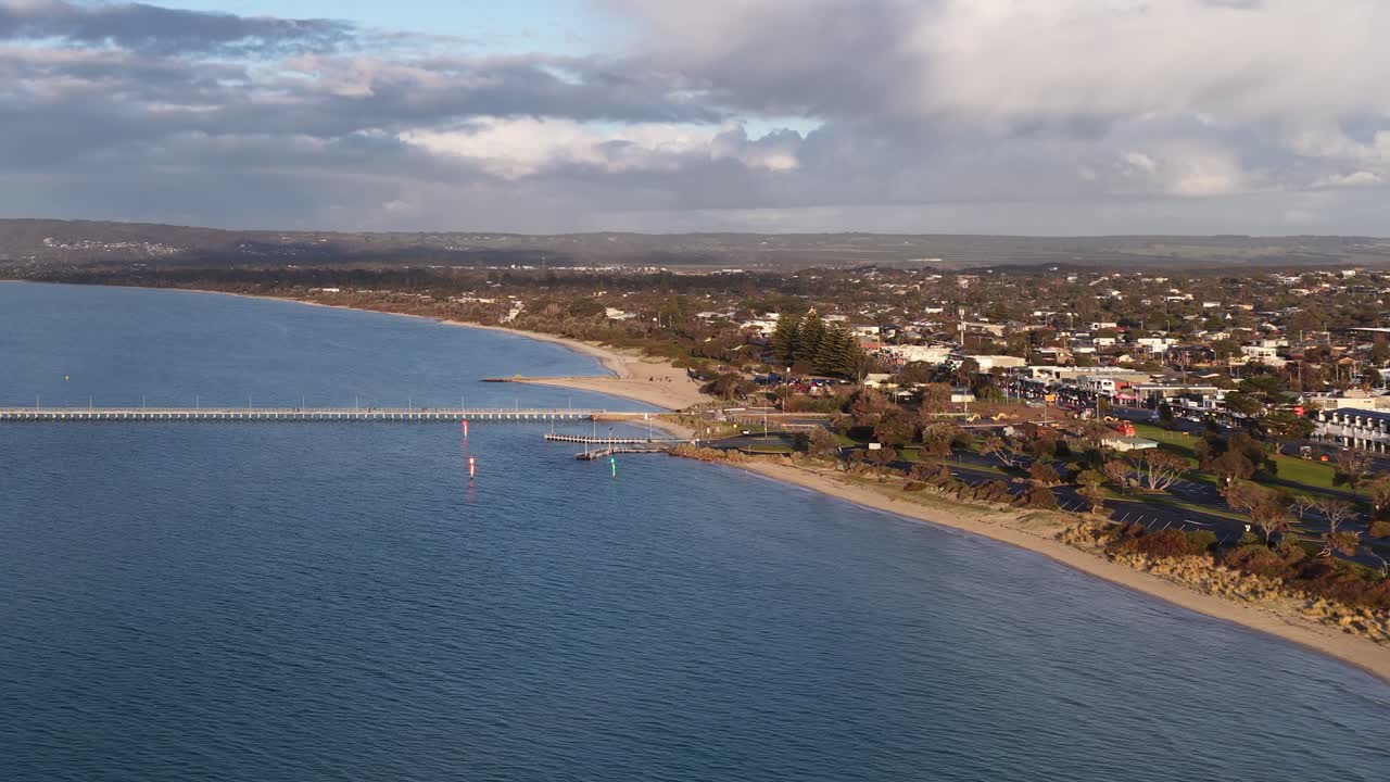 Drone glides above Rye Pier, revealing coastline, calm water, residential area, and soft afternoon light