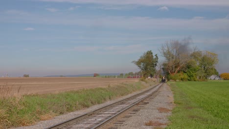 a track side view of a stem passenger train approaching, blowing smoke and steam on a sunny autumn day