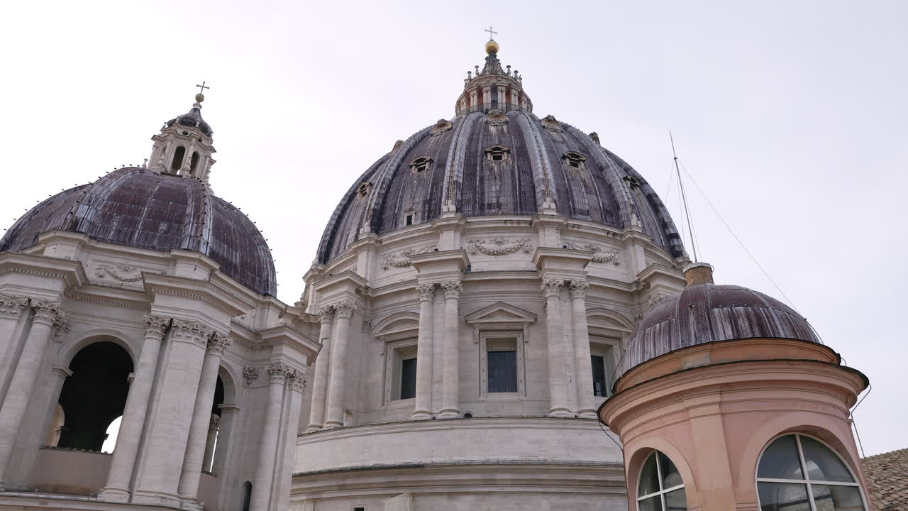 Vista de baixo ângulo da cúpula da Basílica de São Pedro na Cidade do Vaticano, com detalhes arquitetónicos monumentais de travertino contra um céu claro. Símbolo icónico de Roma, religião, arte e viagens.