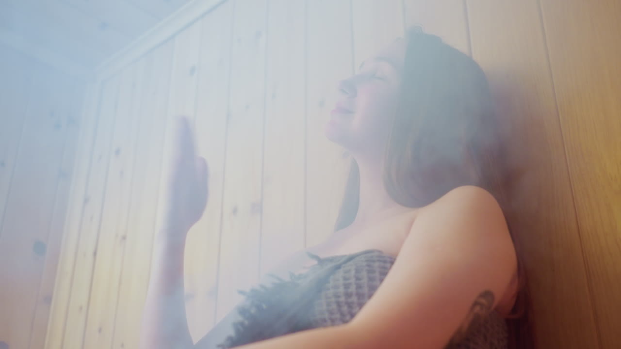 Close up of relaxed girl wrapped in textured towel enjoying steamy moment indoors, resting peacefully against wooden wall with soft warm smile, eyes slightly closed