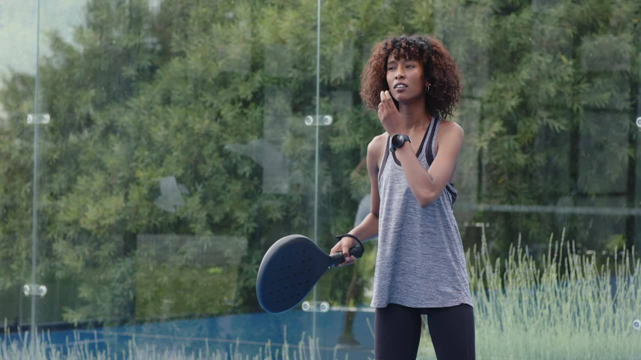 African woman in tank warming up, shifting paddle, checking smartwatch on glass court, copy space