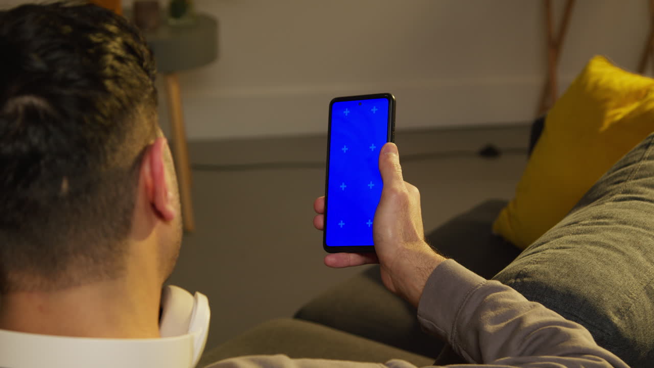 vista trasera de cerca de un hombre pasando la noche en casa sentado en el sofá mirando la pantalla azul del teléfono móvil