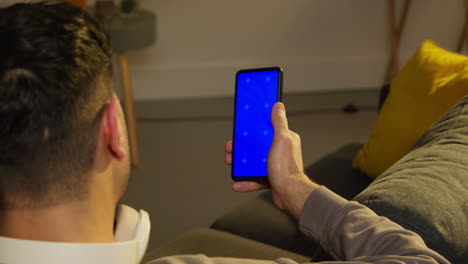 Close Up Rear View Of Man Spending Evening At Home Sitting On Sofa Looking At Blue Screen Mobile Phone