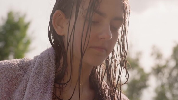 Young woman with wet hair dries herself using pink towel outdoors wearing light purple bathrobe. scene features soft natural lighting and blurred greenery in background focusing on her