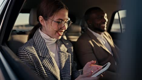 una feliz chica morena con gafas redondas se alegra y se divierte mientras se comunica con su colega, un hombre de piel negra en un traje marrón durante su viaje de negocios en un coche moderno