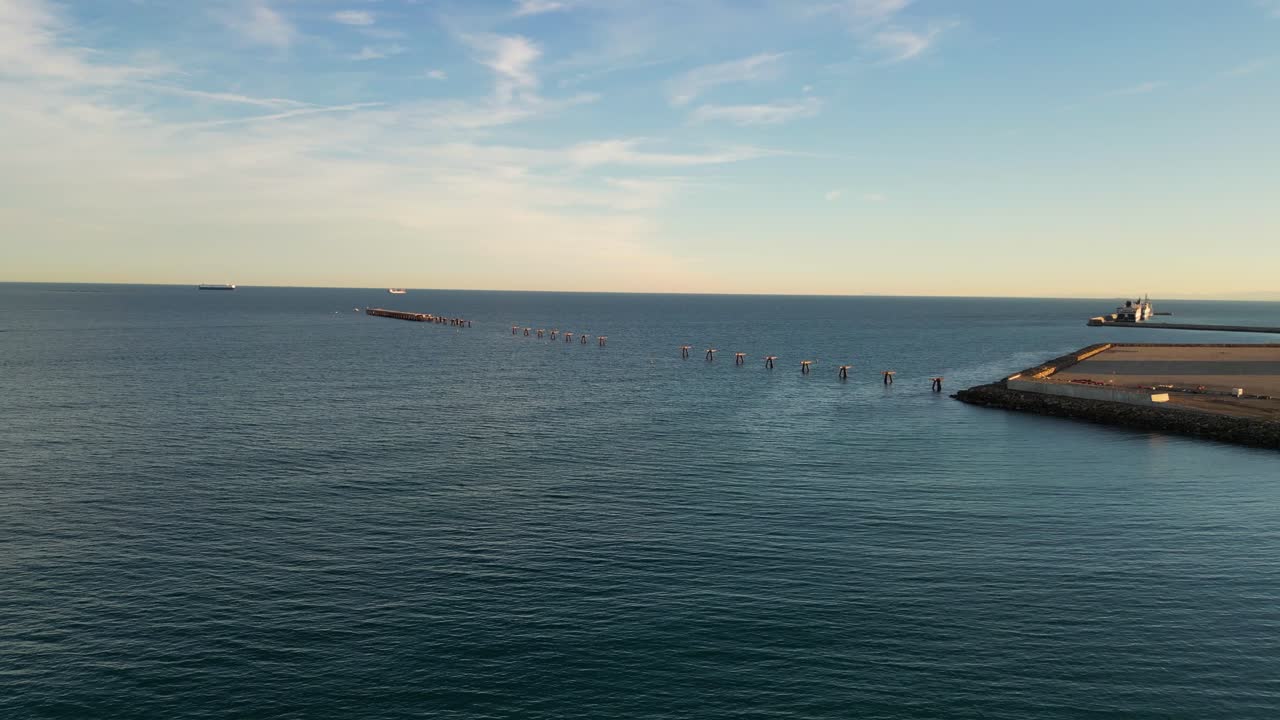 flight in the sea seeing an old pier with the walkway almost gone, only the anchors remaining with boats in the background with a blue sky with clouds on a winter sunset in Valencia Spain