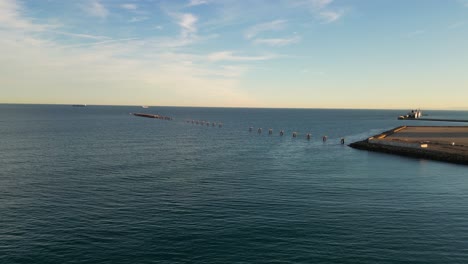 flight-in-the-sea-seeing-an-old-pier-with-the-walkway-almost-gone,-only-the-anchors-remaining-with-boats-in-the-background-with-a-blue-sky-with-clouds-on-a-winter-sunset-in-Valencia-Spain