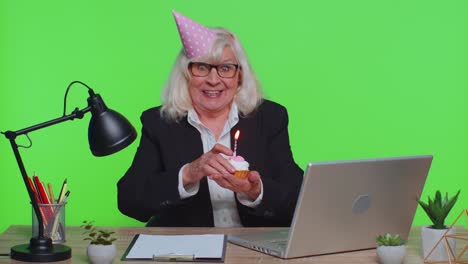 una mujer de negocios senior celebra un cumpleaños solitario en la oficina soplando velas en un pastel pequeño hacer un deseo