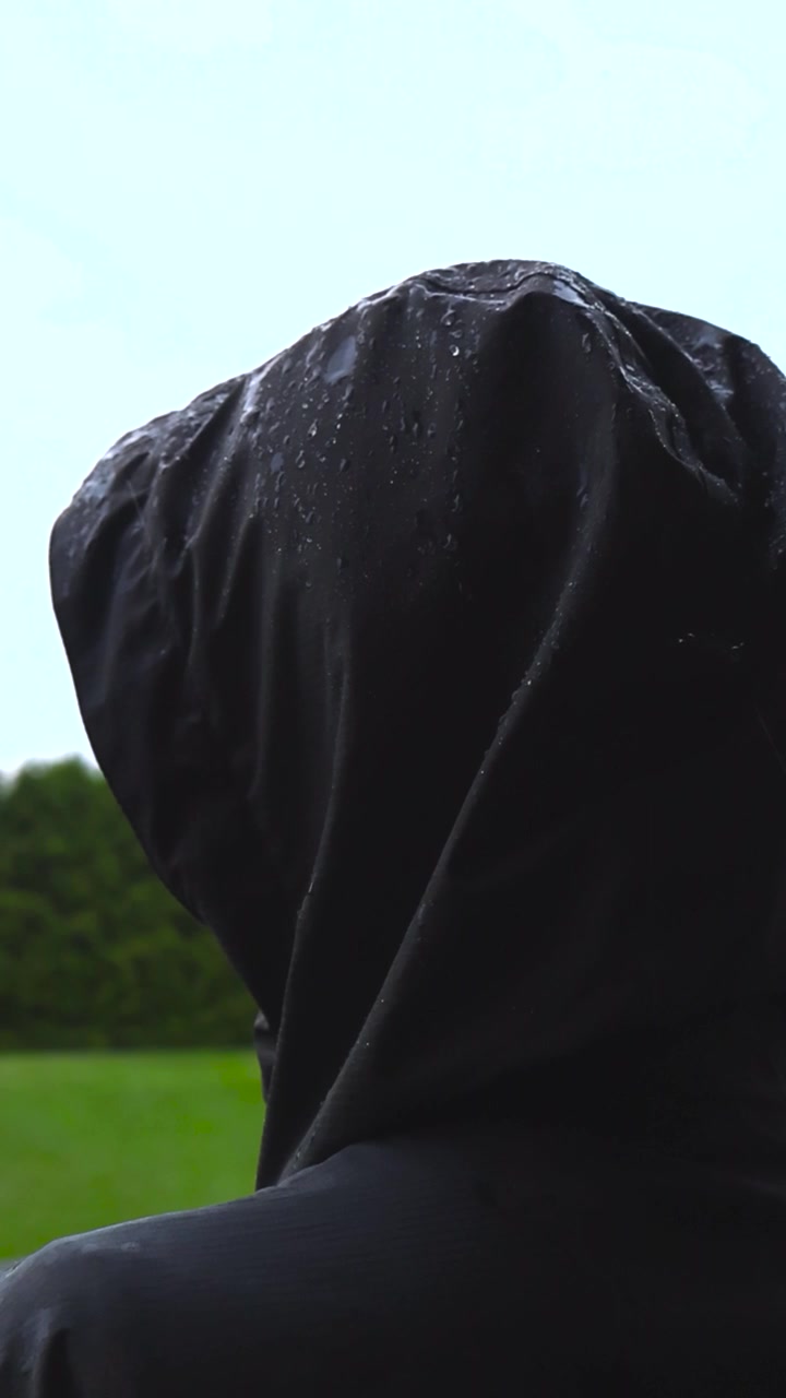 Dramatisk bakfra-utsikt av en anonym person med en svart vannavstøtende hette mens den står i regnet, med vanndråper som faller på stoffet i ekstrem sakte film
