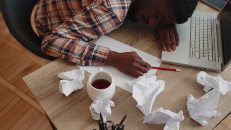 tired man at home office falling asleep on table with laptop computer, crumpled sheets of paper