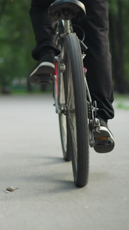 rear view of person in sneakers standing while riding bicycle on paved road, captured from low angle, in the background, there's a blurred view of another person sitting on a bench