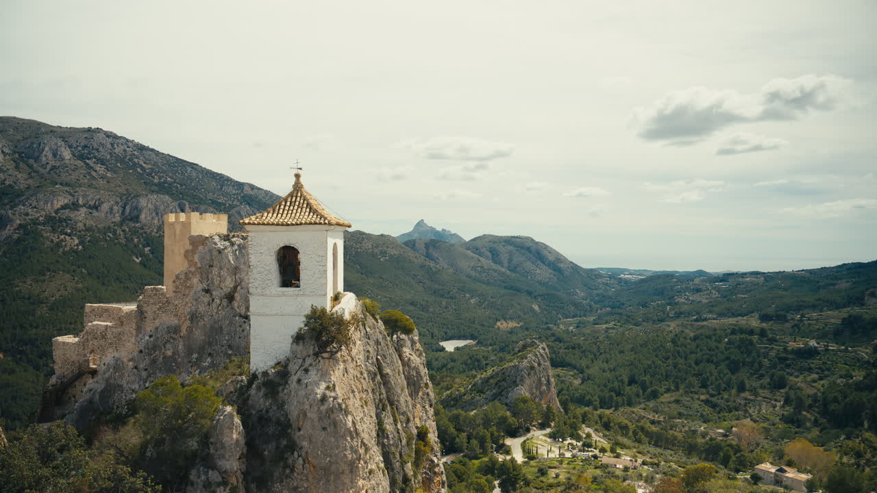 Campanile storico e fortezza arroccati su una drammatica scogliera rocciosa che domina una vasta valle montuosa verde. Vista panoramica elevata con paesaggio esteso e luce naturale soffusa