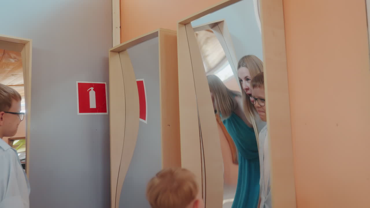 Niños de la familia asomándose a una exhibición de espejos, madre inclinada sobre los niños mientras exploran paneles de reflexión ondulados en el interior, sorpresa juguetona y risas, luz diurna cálida, marcos de madera y colores suaves, casual.