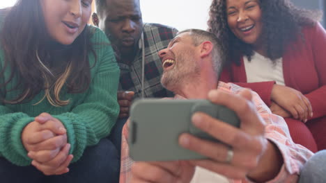 Happy-diverse-male-and-female-friends-relaxing-at-home-together-talking-and-looking-at-smartphone