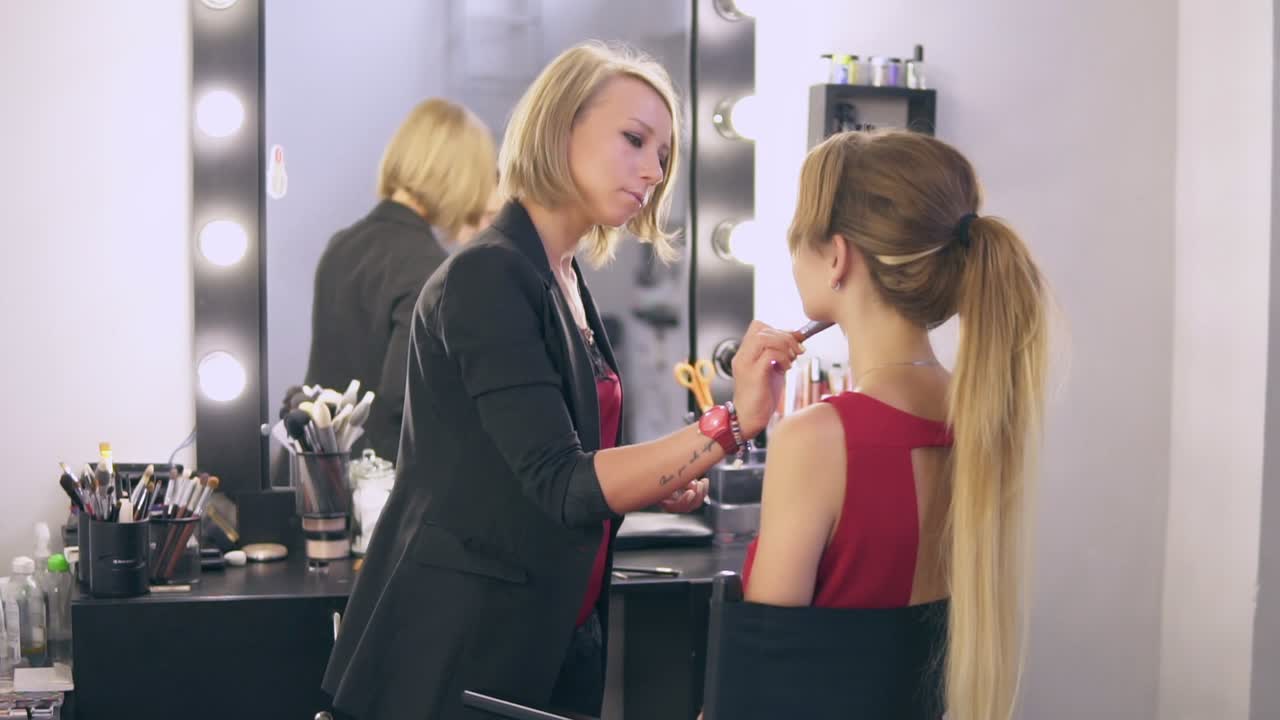 Make-up artist using big brush to apply face powder and finishing make up for a young blonde woman in red dress. Beauty salon