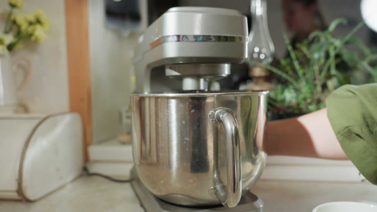 Chef turns off electric mixer after use in home kitchen as stainless bowl rests on mixer base, reflection of chef seen faintly in mirror, surrounded by plants and utensils in bright natural lighting