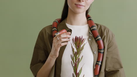 portrait of a young woman with a pet snake around her neck holding a twig on a green background