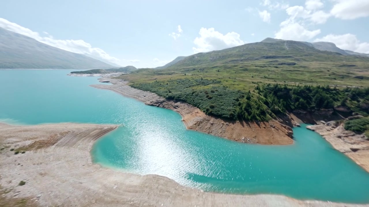 Lago alpino azul-turquesa brilhante margeia terraços rochosos e colinas verdes enquanto a câmera sobrevoa a linha costeira em um dia claro