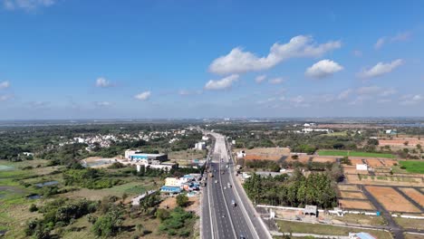 aerial view of a multi-lane highway bustling with traffic, cutting through chennai's diverse landscape of residential areas, fields, and new developments, under a bright sky with scattered clouds